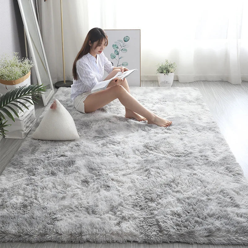 A woman sits on a large, rectangular, fluffy gray plush rug in a bright, modern room.