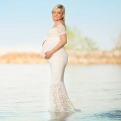 A pregnant woman wears a white off-the-shoulder lace mermaid maternity gown in the water.