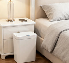 A white rectangular smart trash can with an automatic sensor lid sits beside a bedroom nightstand.