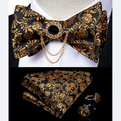 A black and gold paisley patterned bow tie set with a decorative brooch, pocket square, and cufflinks.