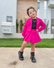 A toddler girl wears a bright pink button-front jacket and matching pleated skirt set.