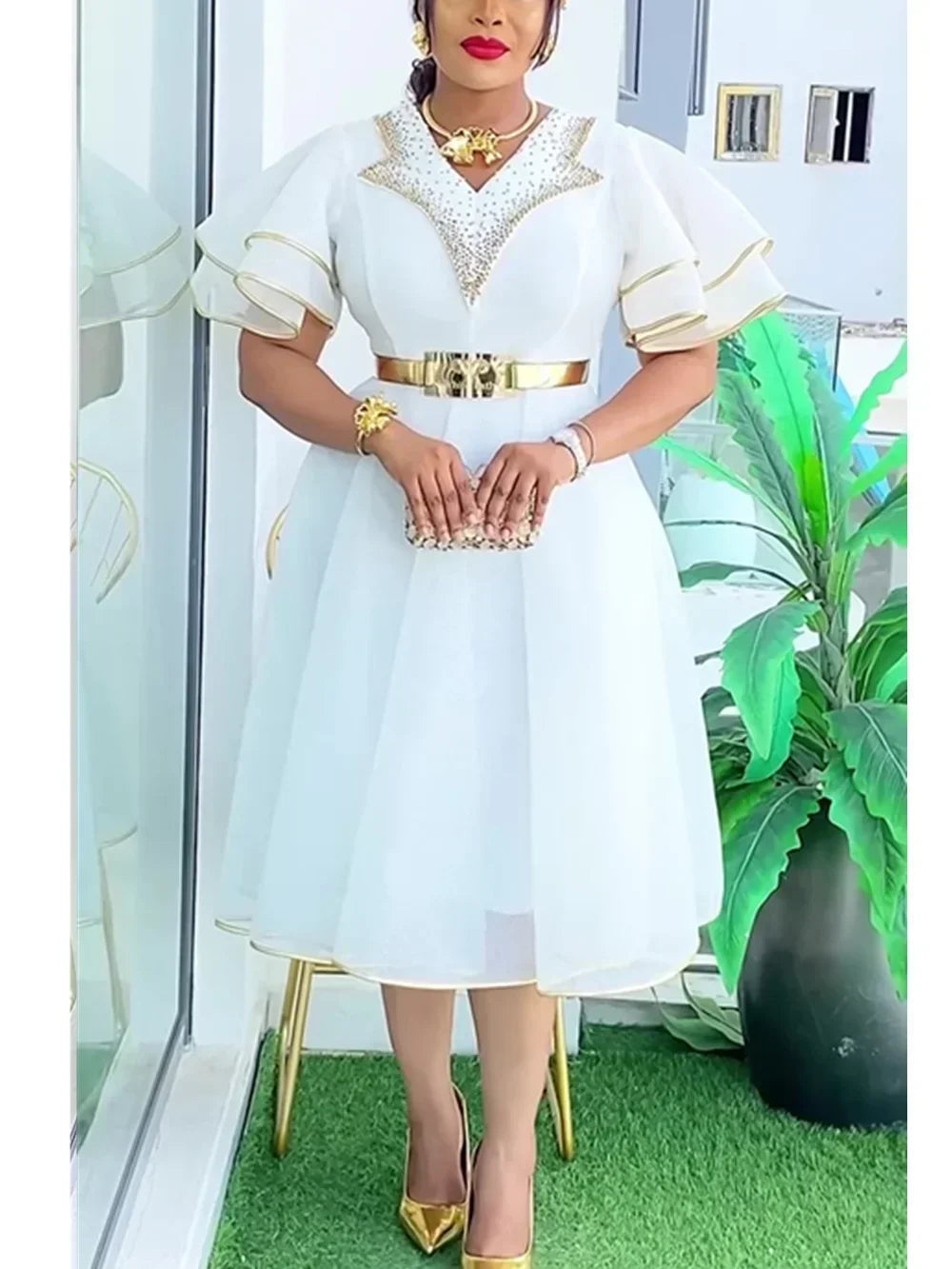 A woman wears a white midi dress with gold trim, a metallic belt, and embellished V-neckline.