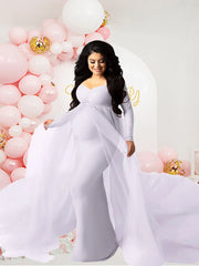 A pregnant woman wears a long-sleeved white floor-length maternity dress with a sheer mesh train.