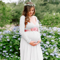 A pregnant woman wears a white long-sleeve lace V-neck maxi dress with a pink floral sash.