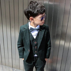 A young boy wears a dark green four-piece formal suit with a white shirt and patterned bow tie.
