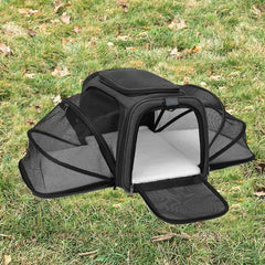 A black foldable pet carrier with mesh sides and a soft interior pad sitting on grass.