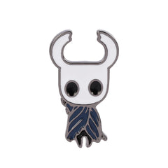 A metal enamel pin of the Hollow Knight character with a white head and blue cloak.
