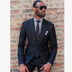 A man wears a black two-piece suit with a black and white gingham shirt and a black tie.