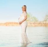 A pregnant woman wears a white off-the-shoulder lace maternity gown while standing in the water.
