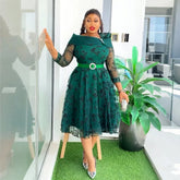 A woman wears a knee-length, dark green dress with black floral embroidery and a matching belt.