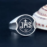 A sterling silver ring featuring the JHS monogram with a cross on a textured black background.