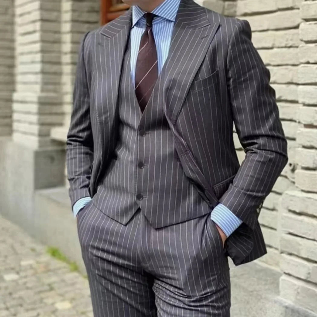 A man wears a charcoal grey pinstripe three-piece suit with a peak lapel and light blue shirt.