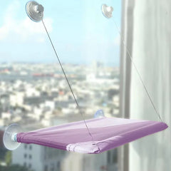A purple cat hammock hangs on a window using four suction cups and support wires.