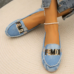A pair of light blue denim loafers featuring a decorative gold-tone logo plate with rhinestones.