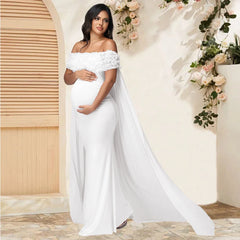 A pregnant woman wears a white off-shoulder maternity maxi dress with floral details and a mesh cape.