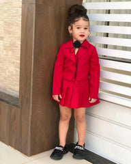 A young girl wears a red long-sleeved blazer and pleated miniskirt set with a black neck accessory.