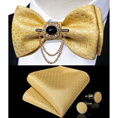 A gold patterned bow tie with a black brooch and chain, matching pocket square, and cufflinks.