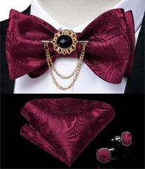 A burgundy paisley bow tie set featuring a decorative brooch with a chain, pocket square, and cufflinks.