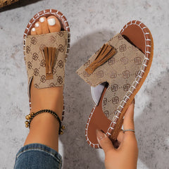 Tan canvas slides featuring a monogram pattern, tassel detail, and white stitched trim.