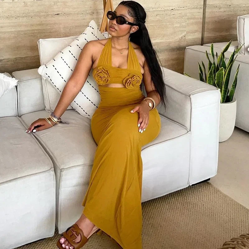 A woman wears a mustard yellow ankle-length halter dress featuring two floral accents on the bodice.