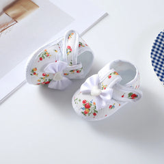 A pair of white baby girl shoes featuring a floral print, a white bow, and a soft sole.