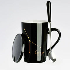 A black ceramic mug featuring a gold Cancer constellation design with a matching lid and spoon.