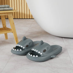A pair of grey shark-shaped EVA slides with open toes sitting on a tiled bathroom floor.
