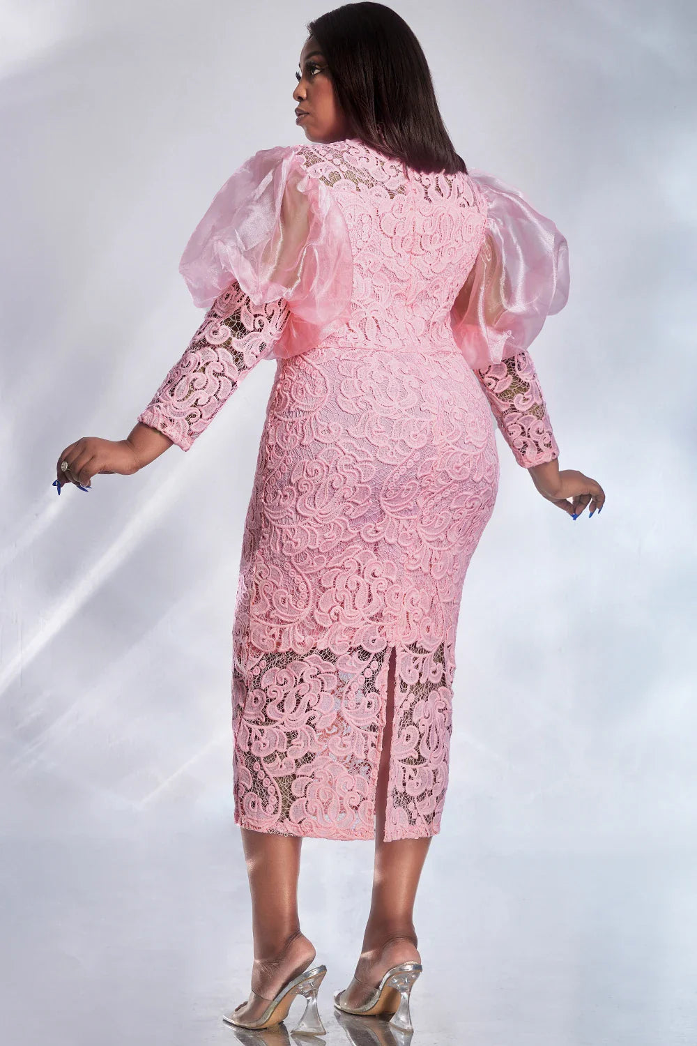 A woman wears a pink lace midi dress with sheer puff sleeves and a back slit.