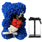 A blue artificial rose bear holding a small bouquet of orange flowers next to a clear gift box.