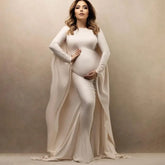 A pregnant woman wears a long-sleeved, ankle-length cream maternity dress with a cape-style back.