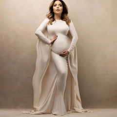 A pregnant woman wears a long-sleeved, ankle-length cream maternity dress with a cape-style back.
