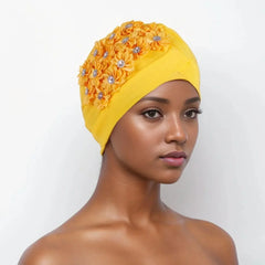 A model wears a bright yellow pleated turban cap decorated with floral accents and rhinestones.