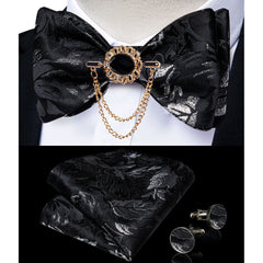 A black and silver paisley bow tie set featuring a decorative brooch, pocket square, and cufflinks.