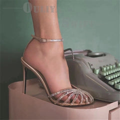 A foot wearing a champagne-colored stiletto sandal with crystal-embellished straps and a buckle.