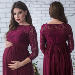 A pregnant woman wears a burgundy lace maternity maxi dress with three-quarter sleeves and a sash.