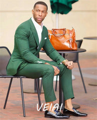 A man wears a tailored green two-piece suit with a white shirt and black loafers.