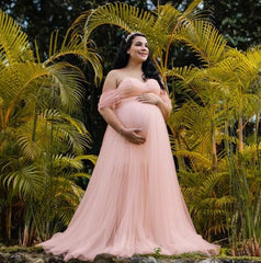 A pregnant woman wears a pink off-shoulder chiffon maxi dress for a photoshoot.