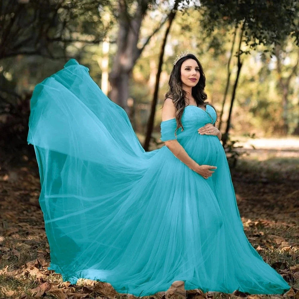 A pregnant woman wears a flowing blue off-shoulder maternity gown in a wooded setting.