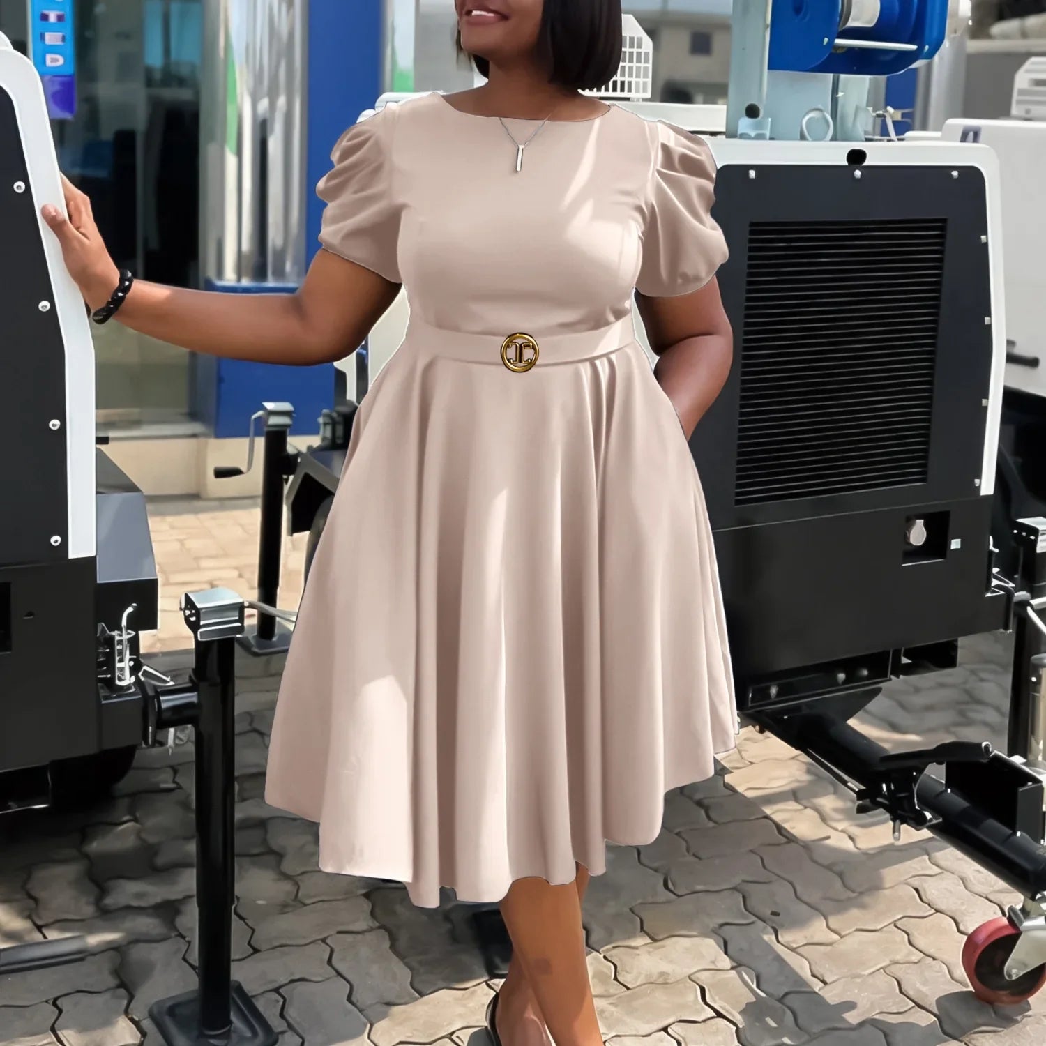 A woman wears a beige, short-sleeved, pleated midi dress with a gold-buckled belt.