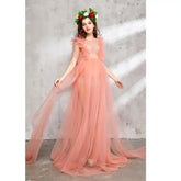 A pregnant woman wears a sleeveless, ankle-length peach tulle maternity dress with a floral crown.