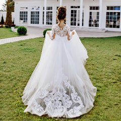 A girl wears a floor-length white lace flower girl dress with long sleeves and a train.