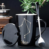 A black ceramic mug featuring a gold Libra constellation design, complete with a lid and spoon.