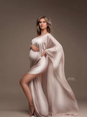 A pregnant woman wears a strapless, ankle-length satin maternity cape in a soft beige tone.