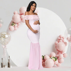 A pregnant woman wears a light pink off-shoulder maternity gown with floral details and chiffon hem.