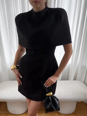 A woman wears a black mini dress featuring a high collar and a structured shawl overlay.