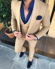 A man wears a slim-fit tan three-piece suit with a blue shirt and matching navy loafers.