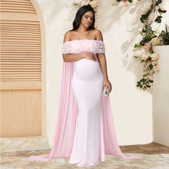 A pregnant woman wears a light pink off-shoulder maxi dress with floral detailing and a mesh cloak.