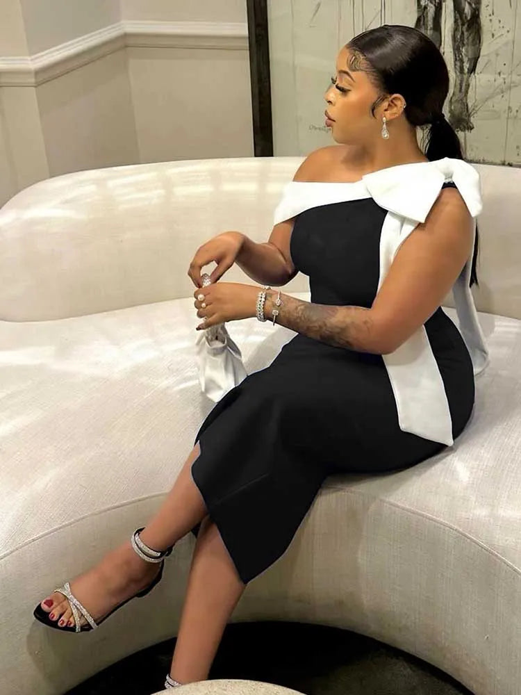 A woman wears a black mid-length dress featuring a white bow detail on the shoulder.