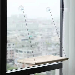 A beige cat hammock hangs from a window using four suction cups and thin support cables.