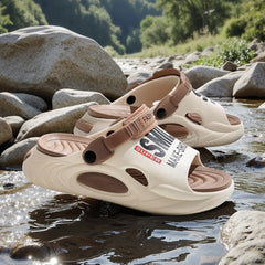 Beige and brown EVA slides with thick soles and letter detailing resting on river rocks.
